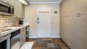 Una cocina compacta equipada con lavavajillas, microondas y lavabo. Hay una puerta con perchero y un cartel encima. El suelo tiene un diseño de baldosas con motivos.