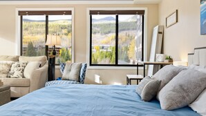Un dormitorio con una cama grande, un sofá y un escritorio. Hay dos ventanas con vistas a árboles y montañas.