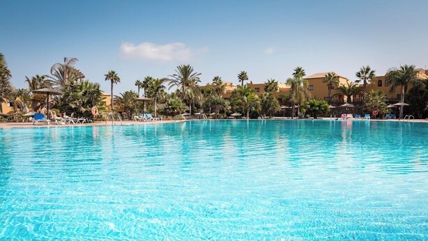 Ein großer, klares Schwimmbecken mit Liegestühlen und Sonnenschirmen, umgeben von Palmen und Gebäuden.