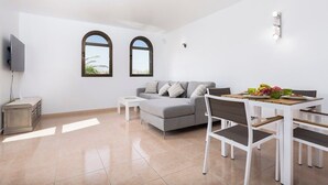 Ein modernes Wohnzimmer mit grauem Sofa, weißem Esstisch und Stühlen, einem Flachbildfernseher und zwei Rundbogenfenstern.