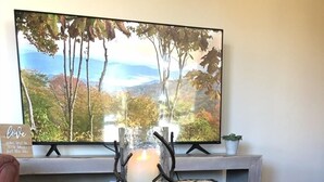 Pyöreä puinen pöytä, jonka keskellä on kynttilänjalka, seinälle asennettu televisio, jossa näkyy kaunis maisema, ja pieni ruukkukasvi sivupöydällä.