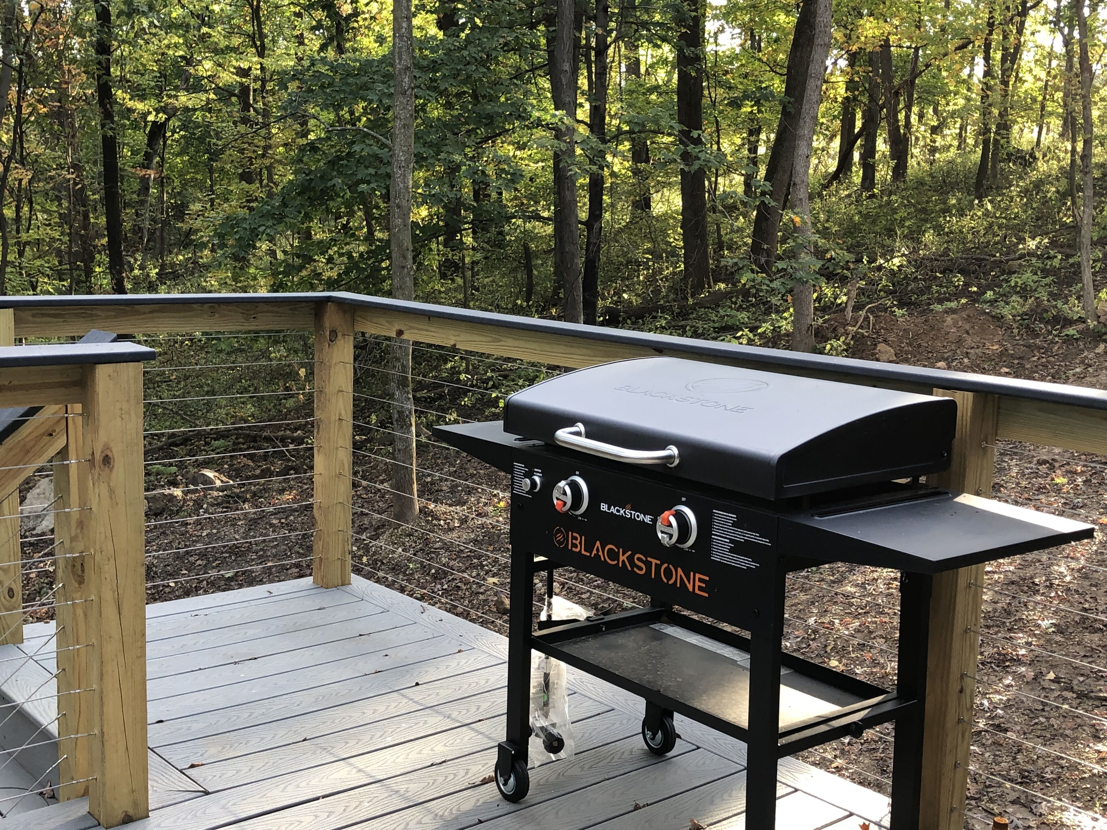 Un barbecue Blackstone portable, installé sur une terrasse en bois dotée d’une rambarde, et entouré d’une zone boisée.