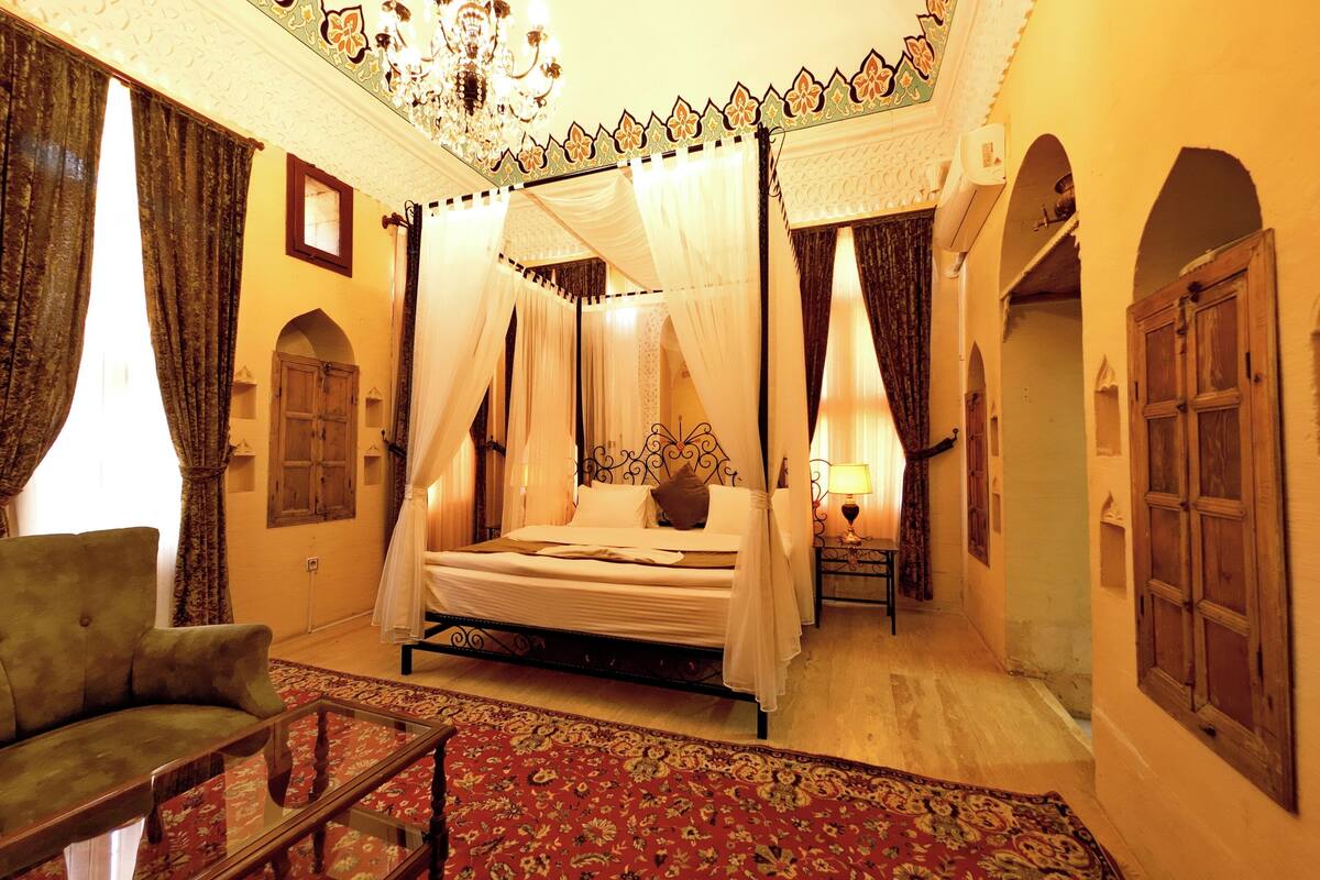 Una habitación tradicional con una cama con dosel, un sillón verde y una alfombra estampada.