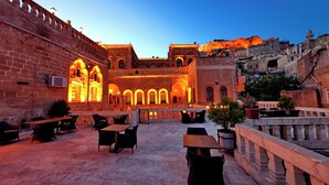 An outdoor café area with stone walls and arched windows, furnished with tables and chairs, and a view of a historic building in the background. - Ulubey Konagi (Mardin)