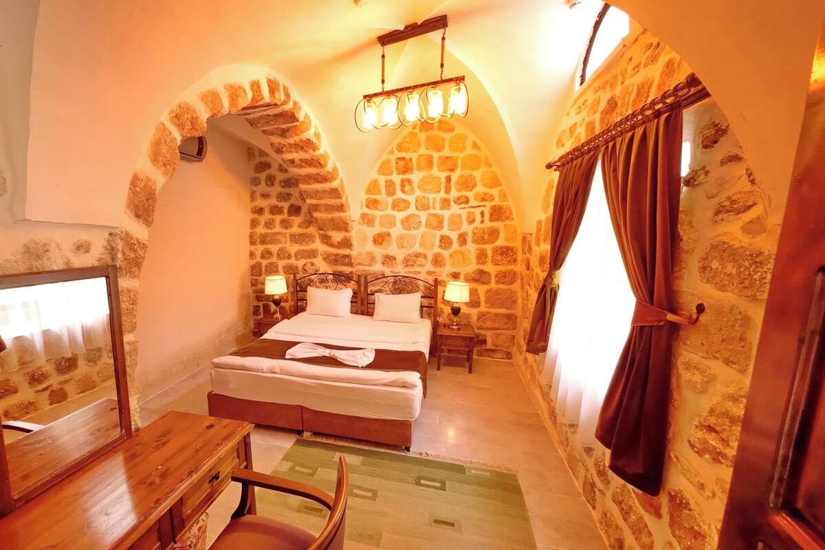 Una habitación con pared de piedra, cama con ropa blanca, dos lámparas de noche y un escritorio de madera.