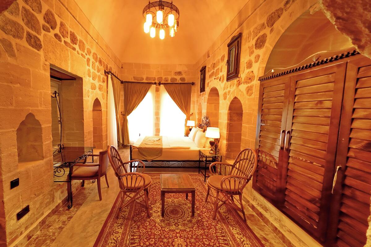 Una habitación con paredes de piedra, una cama con dosel, una mesa, sillas y una araña.