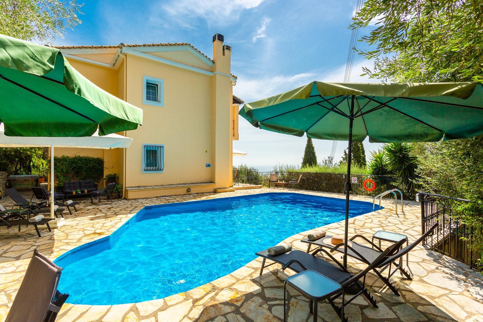 Una piscina amb gandules i para-sols, envoltada per un paviment de pedra i un edifici amb teulada de rajola.