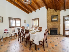 Un coin repas avec une table dressée pour six personnes, des chaises en bois, un plafond haut avec des poutres apparentes, une cheminée en briques et une fenêtre offrant une vue agréable.