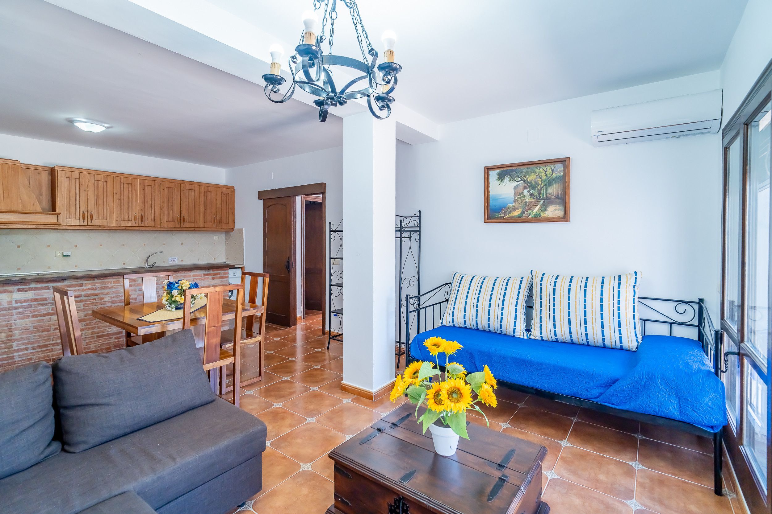 Una sala de estar con sofá, un comedor con mesa y sillas, una cocina con armarios de madera, una araña y un cuadro en la pared.
