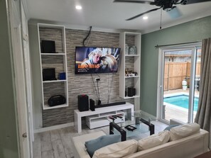 A living room with a flat-screen TV mounted on a wall, a white entertainment unit, a glass coffee table, and a beige sofa. - Brand New, Modern Style 1 BED 1 BATH Cottage, [Gated Compound]. (Nassau)