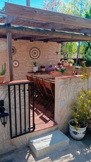 Un patio al aire libre con mesa y sillas de madera, rodeado de plantas en macetas y un muro decorativo.