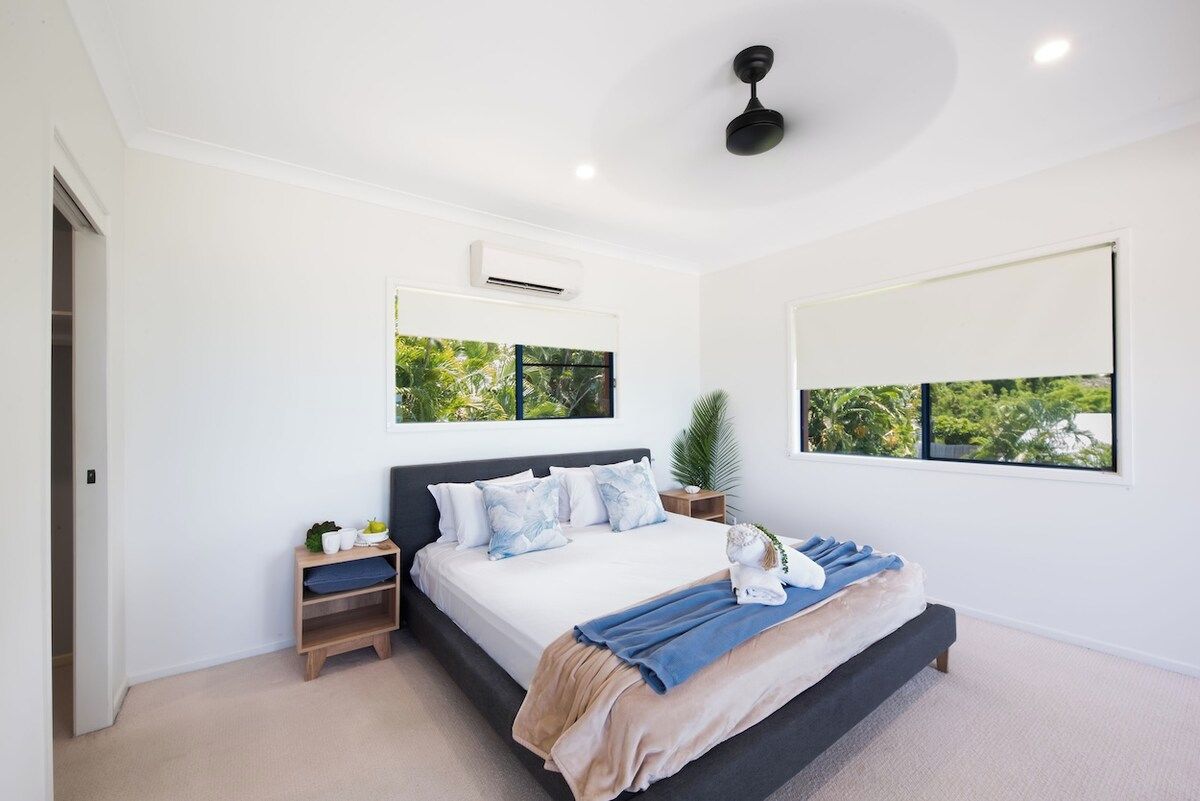 A modern bedroom with a bed, bedside tables, a ceiling fan, and a large window with a view of greenery.