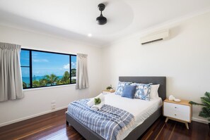 A bedroom with a large window, a bed with patterned bedding, a bedside table, and a ceiling fan. - Beyond The Palms (Airlie Beach)
