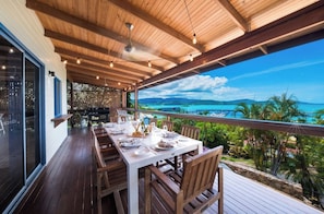 Ein Essbereich im Freien mit einer Holzterrasse, einem Tisch für zwei Personen und Blick auf das Meer und die Berge.