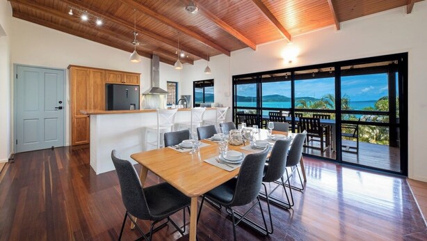 Uma sala de jantar ampla com mesa e cadeiras de madeira, uma cozinha com armários de madeira e grandes janelas com vista para o oceano.