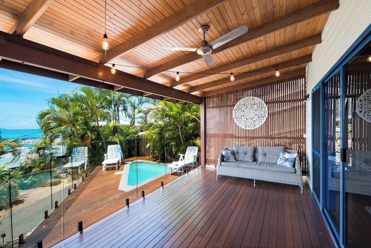 A wooden deck with a pool, lounge chairs, and a sofa with a view of the ocean.