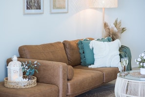 Un sofá de cuero marrón con almohadones decorativos, un farol blanco y una mesita con superficie de vidrio.