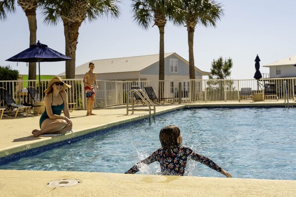 A swimming pool with a child in the water, adults on the edge, and palm trees in the background.