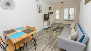 Uma sala de estar moderna com um sofá cinzento, mesa de jantar de madeira com cadeiras, e uma televisão sobre um móvel branco.