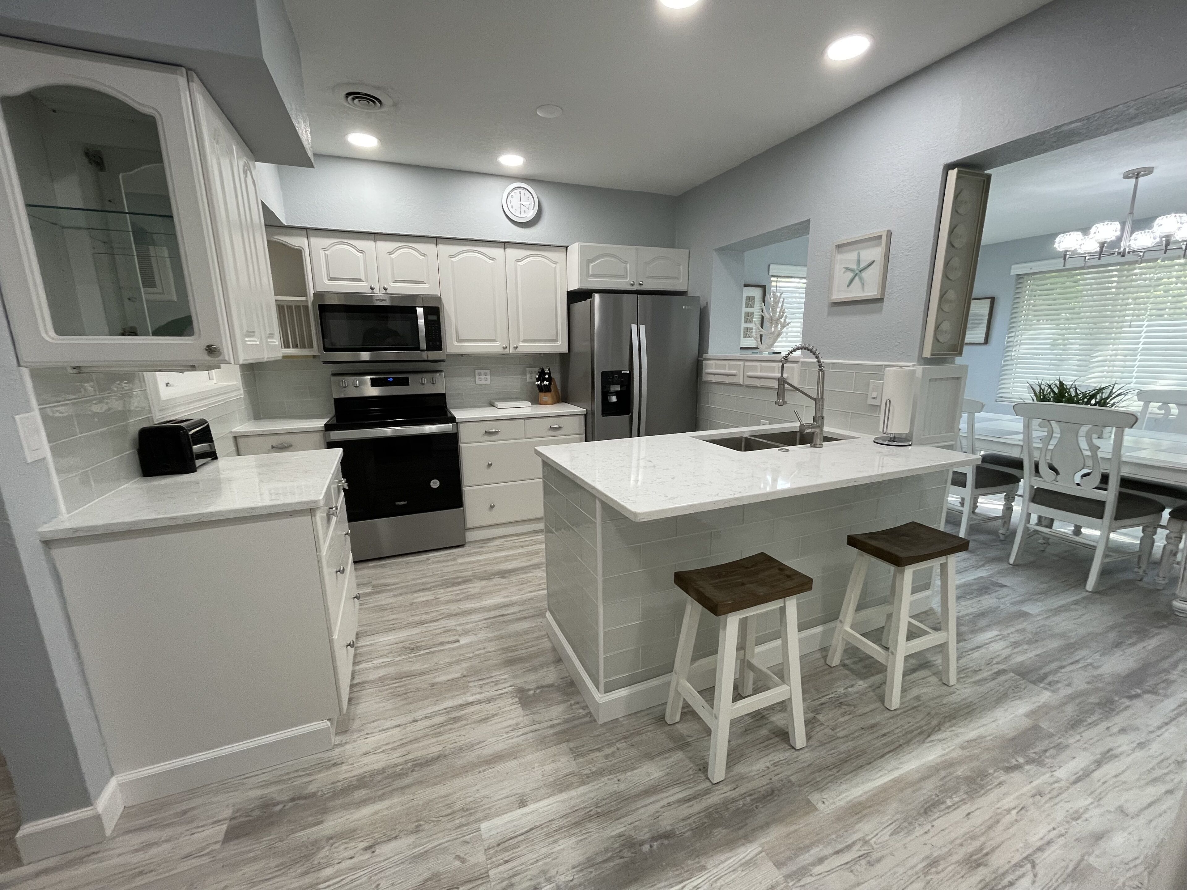 A modern kitchen with white cabinetry, stainless steel appliances, a central island with stools, and a dining area with a white table and chairs.