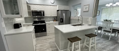 Una cocina moderna con armarios blancos, electrodomésticos de acero inoxidable, una isla central con taburetes y un comedor con mesa y sillas blancas.