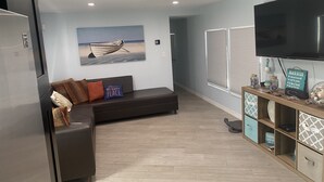 Una sala moderna con un sofá marrón oscuro, un televisor de pantalla plana, un mueble de madera con objetos decorativos y un cuadro de un barco en la pared.