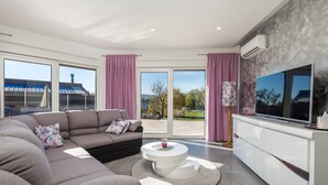 A modern living room with a grey sofa, a white coffee table, and a TV on a white cabinet. Large windows with pink curtains offer a view of a garden and a house. - Gorgeous home in Rijeka (Rijeka)
