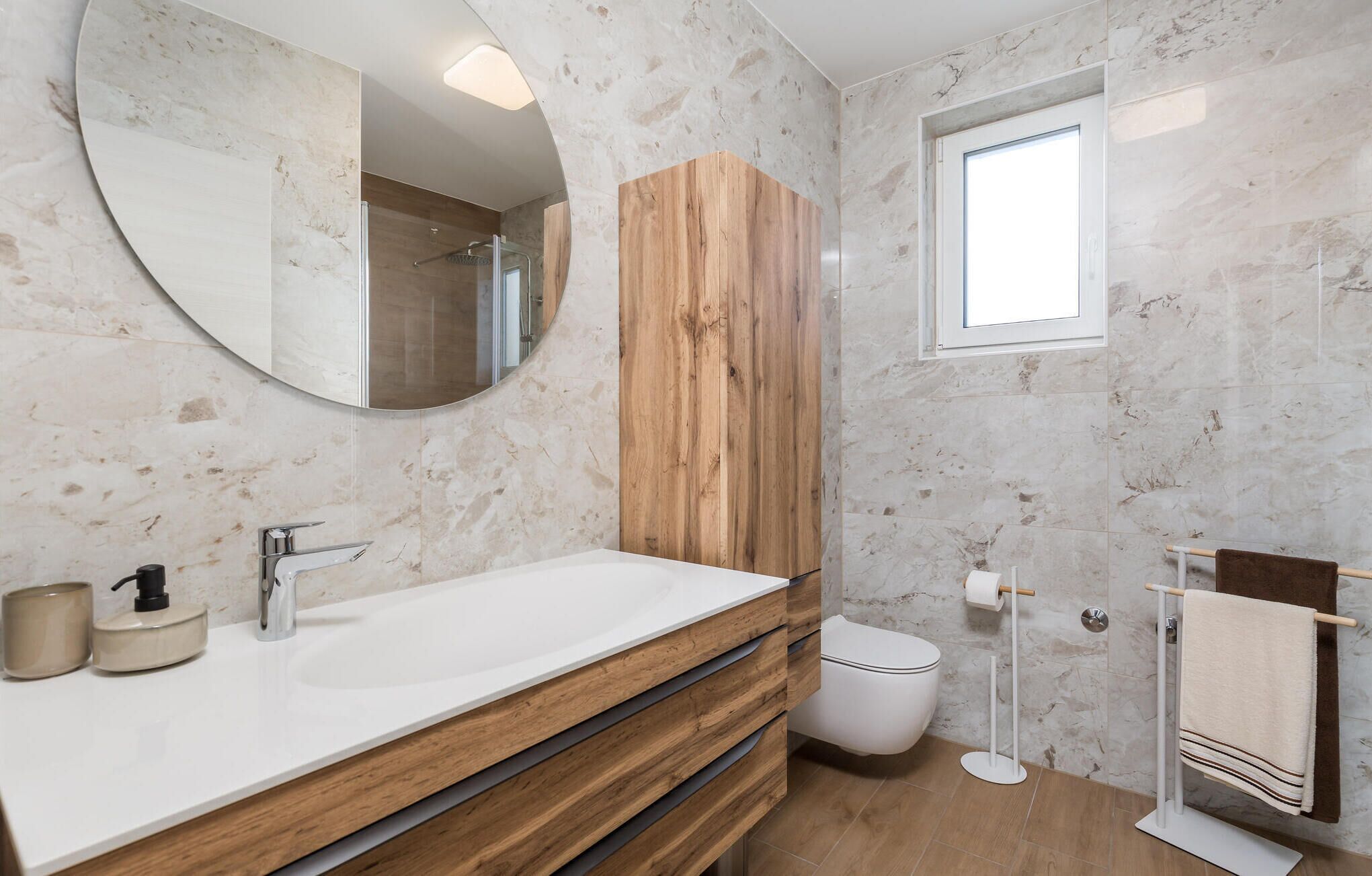 Et badeværelse med en trækommode, et rundt spejl, et toilet og en håndklæderække.