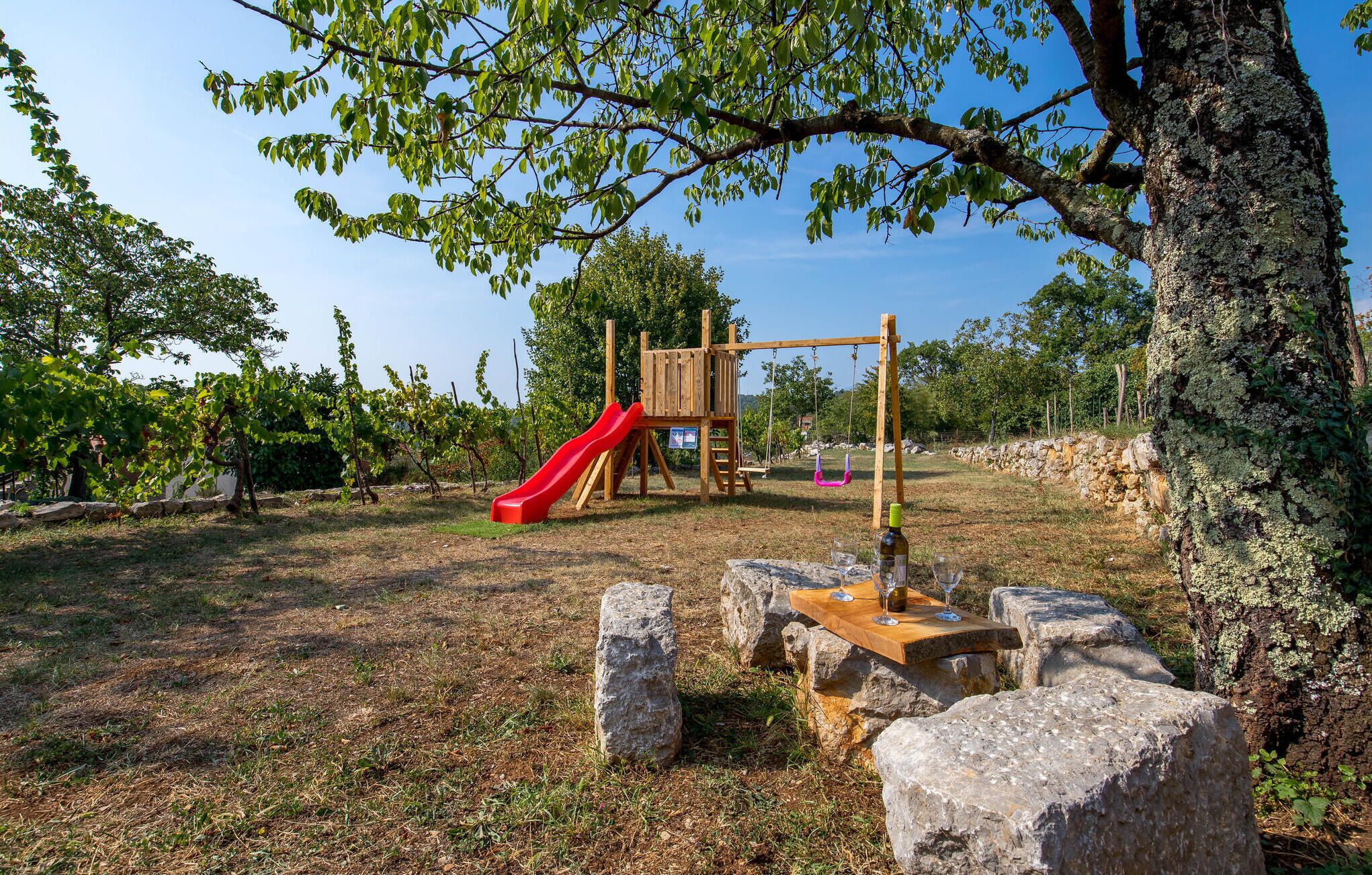 A playground with a red slide and a wooden climbing structure, surrounded by stone seating and a table with wine glasses.