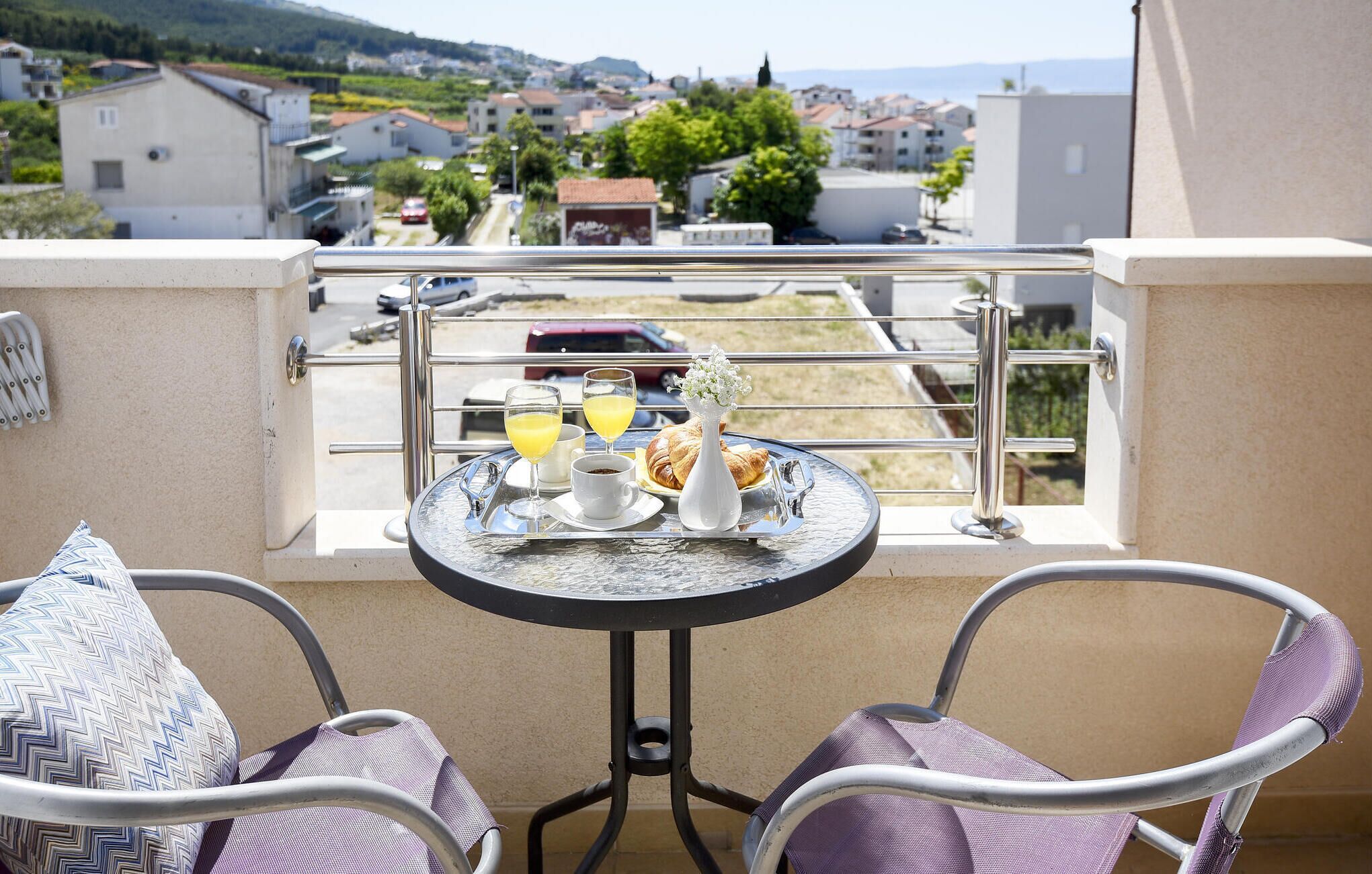 A balcony with a table set for breakfast, including croissants and orange juice, overlooking a residential area.