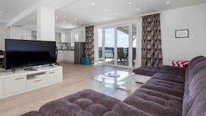 A modern living room with a flat-screen TV, a sofa, a glass coffee table, and a kitchen in the background. - Awesome home in Banjol (Banjol)