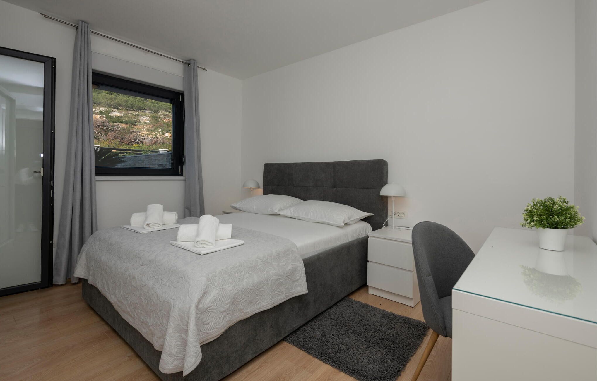 Une chambre moderne avec un lit gris, du linge de lit blanc, une tête de lit grise, une table de chevet avec une lampe, un petit bureau avec une plante, et une vue sur la verdure par la fenêtre.