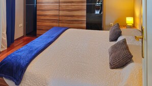 Una habitación de hotel con una cama grande, una manta azul decorativa, dos almohadas decorativas, un armario de madera y una lámpara de noche.