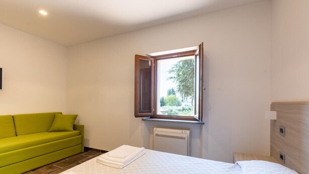 Une chambre d’hôtel avec un lit, un canapé vert, une fenêtre donnant sur un paysage verdoyant et un radiateur.