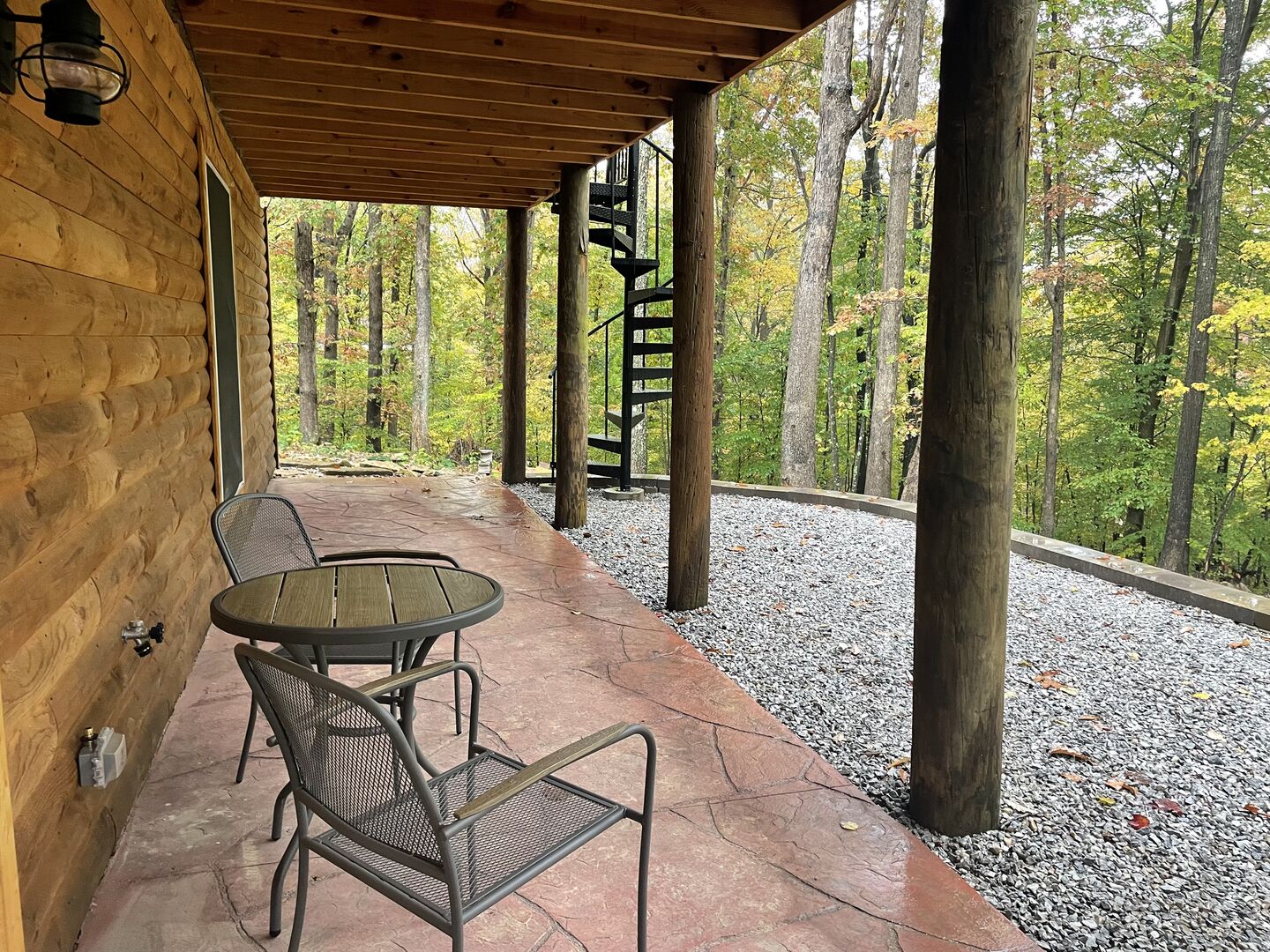 A covered outdoor seating area with a table and chairs, surrounded by trees and a gravel path.