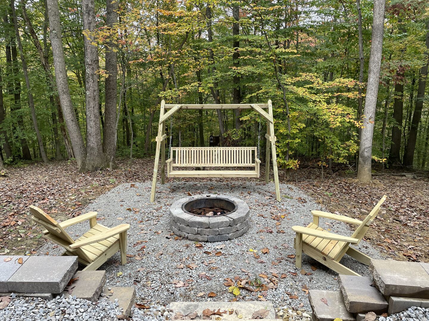 Ein Spielplatz aus Holz mit einer Schaukel, einer Sitzbank, einem Feuerkorb und zwei Holzstühlen in einer bewaldeten Gegend.