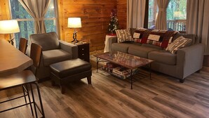 A cozy cabin interior with a sofa, armchair, coffee table, and a small dining table. - Unique Cavalier Log Cabin nestled in the forest, 15 mins to Hocking Hills (Logan)
