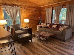 A cozy cabin interior with a sofa, armchair, coffee table, and a small dining table. - Unique Cavalier Log Cabin nestled in the forest, 15 mins to Hocking Hills (Logan)