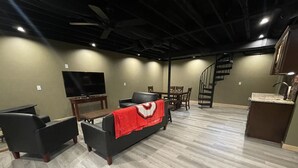 A modern living room with a flat-screen TV, a sofa with a red blanket, a dining table, and a staircase leading to an upper level. - Unique Cavalier Log Cabin nestled in the forest, 15 mins to Hocking Hills (Logan)