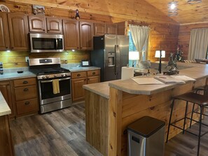 A kitchen with wooden cabinets, stainless steel appliances, a central island, and a bar stool. - Unique Cavalier Log Cabin nestled in the forest, 15 mins to Hocking Hills (Logan)
