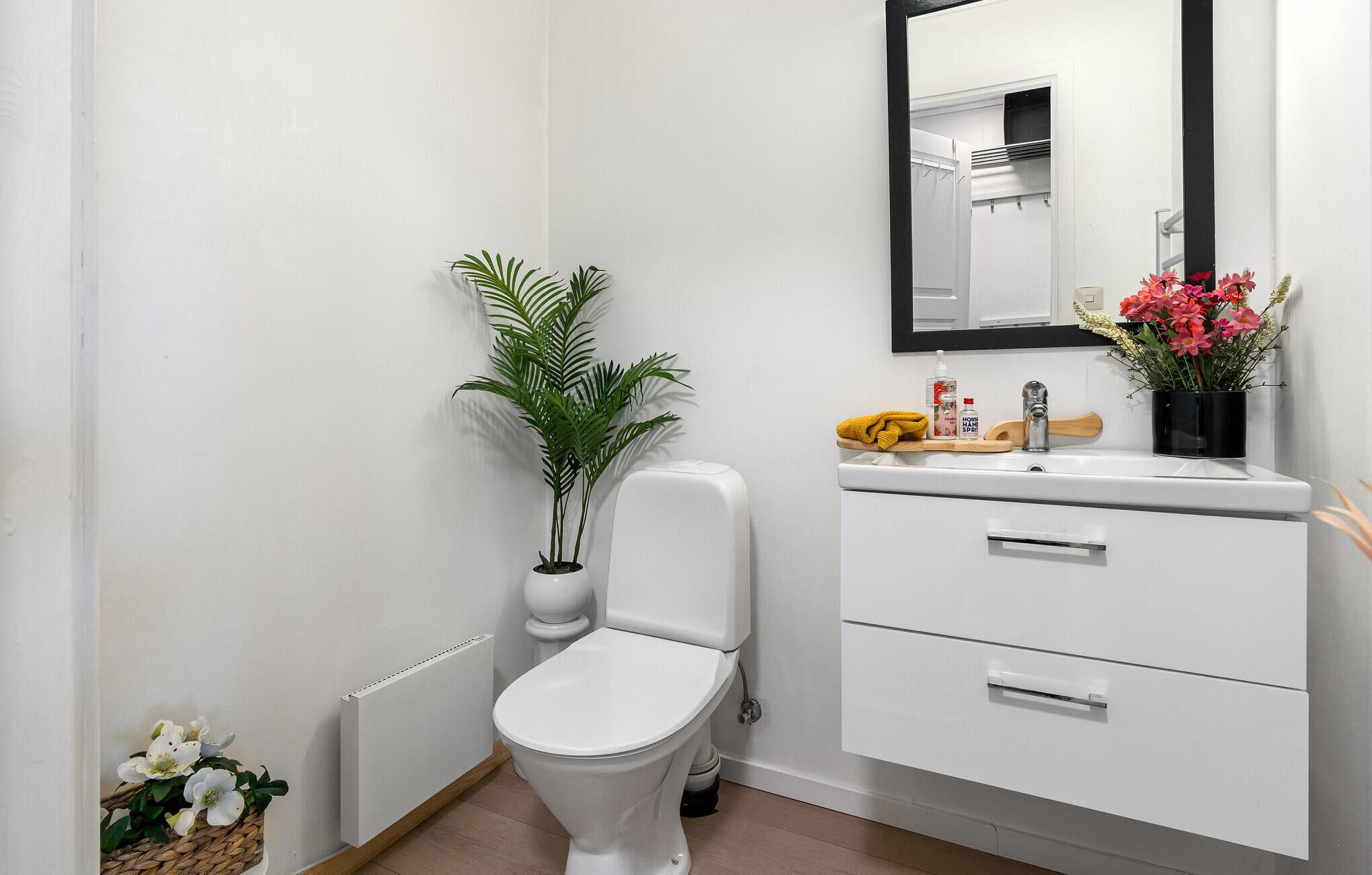 Un baño moderno con inodoro, un mueble blanco con cajones, un espejo, un radiador, una planta en maceta y una pequeña cesta con flores blancas.