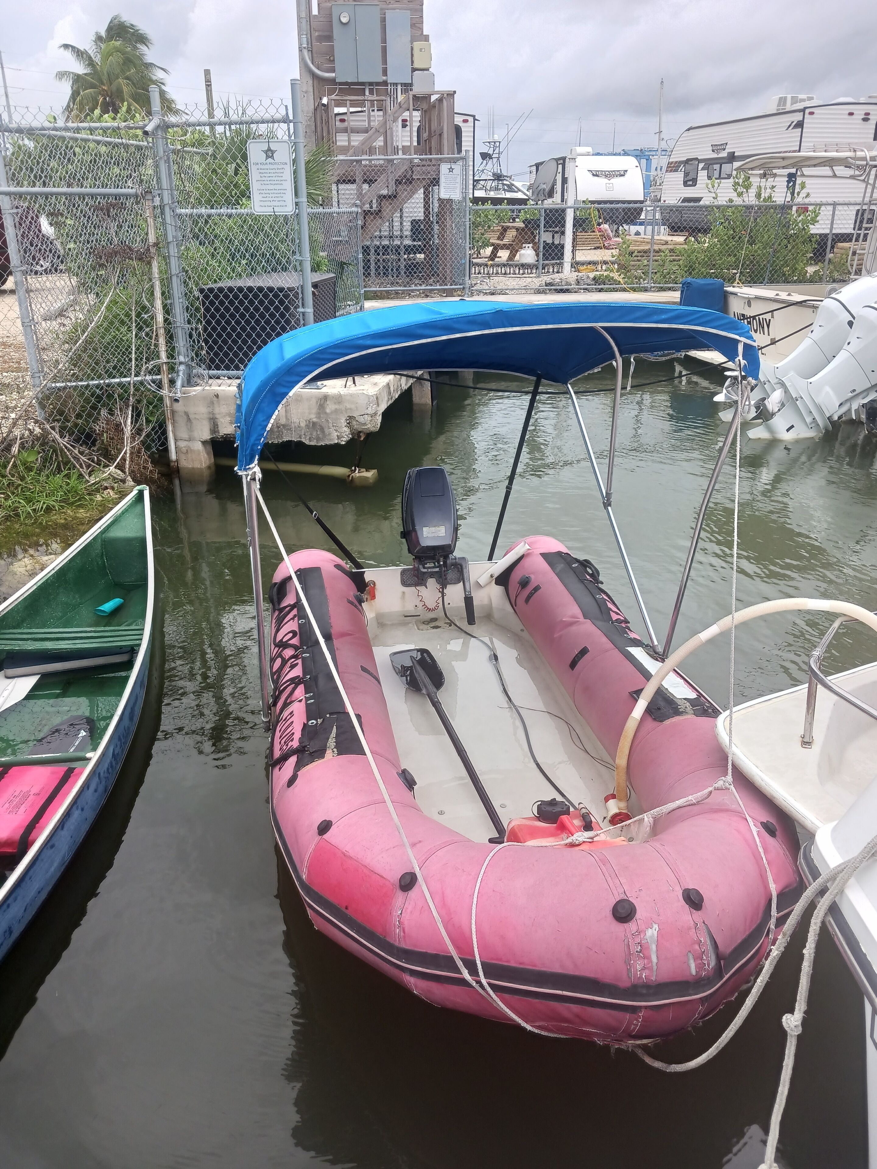 A pink inflatable boat docked at a marina with a blue canopy, surrounded by other boats and a chain-link fence.