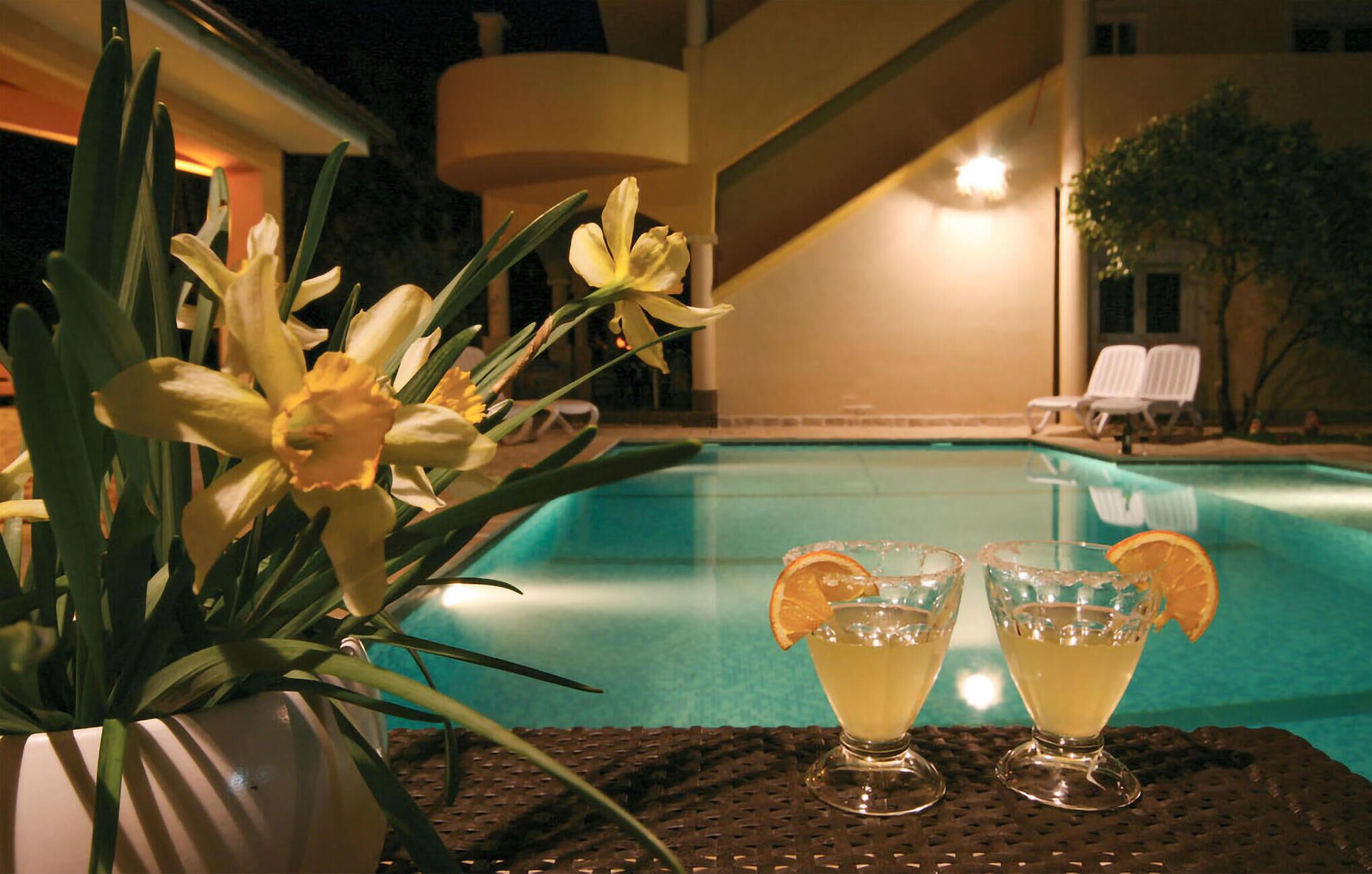 Une scène au bord de la piscine, avec une table sur laquelle reposent deux boissons décorées de tranches d’orange, le tout entouré de fleurs jaunes.
