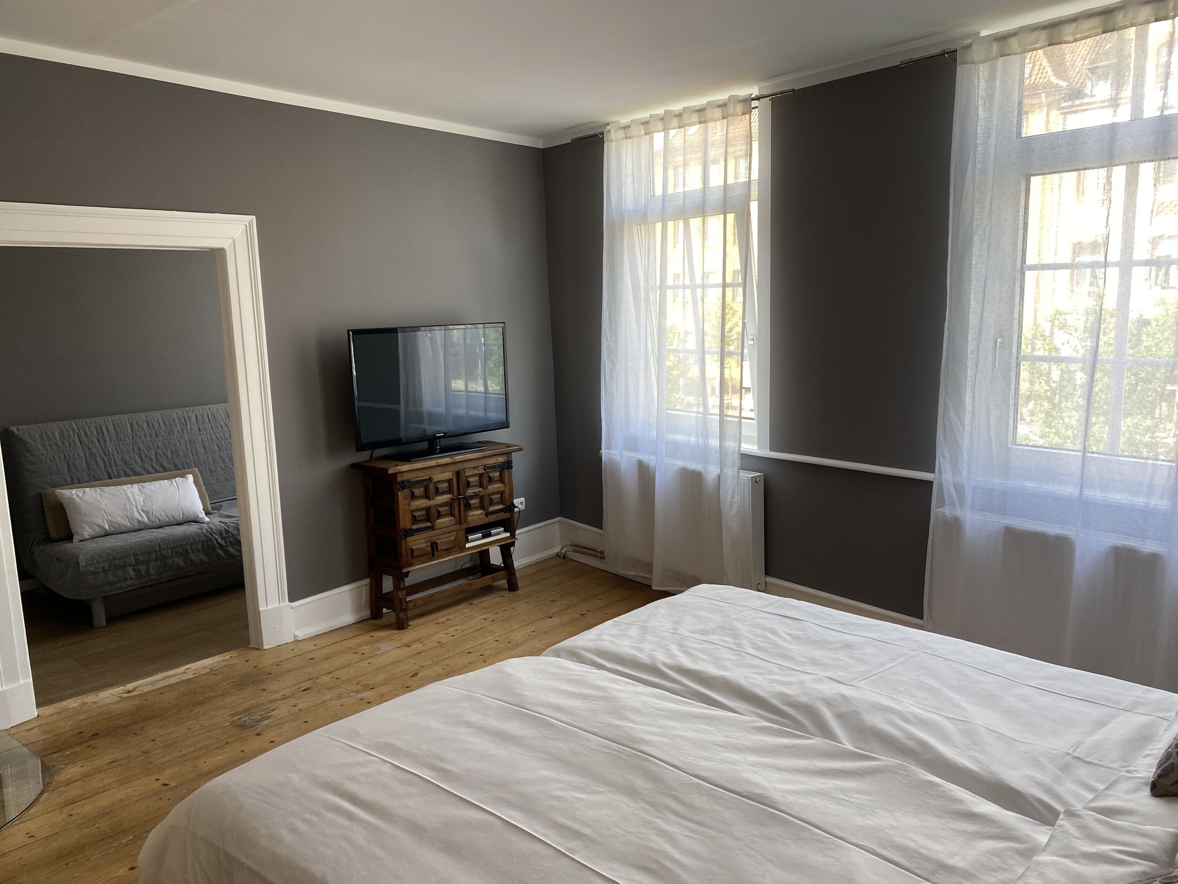 Une chambre à coucher avec un lit, un téléviseur posé sur une commode en bois et une fenêtre ornée de rideaux légers.