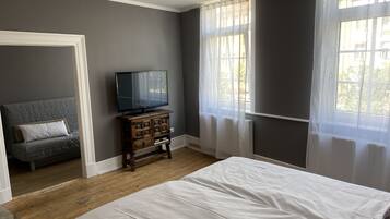 Une chambre à coucher avec un lit, un téléviseur posé sur une commode en bois et une fenêtre ornée de rideaux légers.