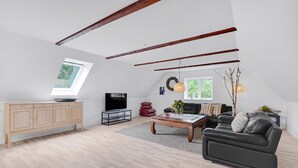 Ein geräumiger Loft mit hoher Decke, freiliegenden Holzbalken, einem großen Fenster, einem Flachbildfernseher, einem Ledersofa und einem Holzschrank.