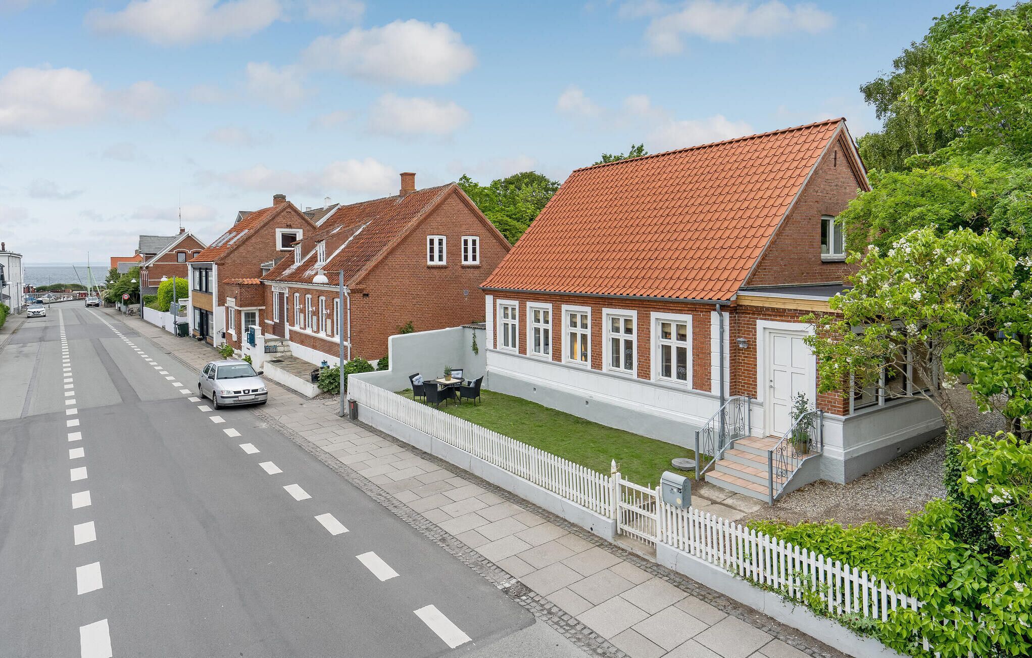Une rangée de maisons en briques avec des toits couverts de tuiles rouges, une clôture blanche en bois et une route dégagée où l’on aperçoit une voiture.