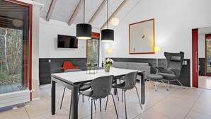 A modern dining area with a white table, black chairs, and a grey sofa. There is a TV mounted on the wall, a large framed picture, and a window with a view of trees. - Amazing home in Grenaa with kitchen (Grenaa)