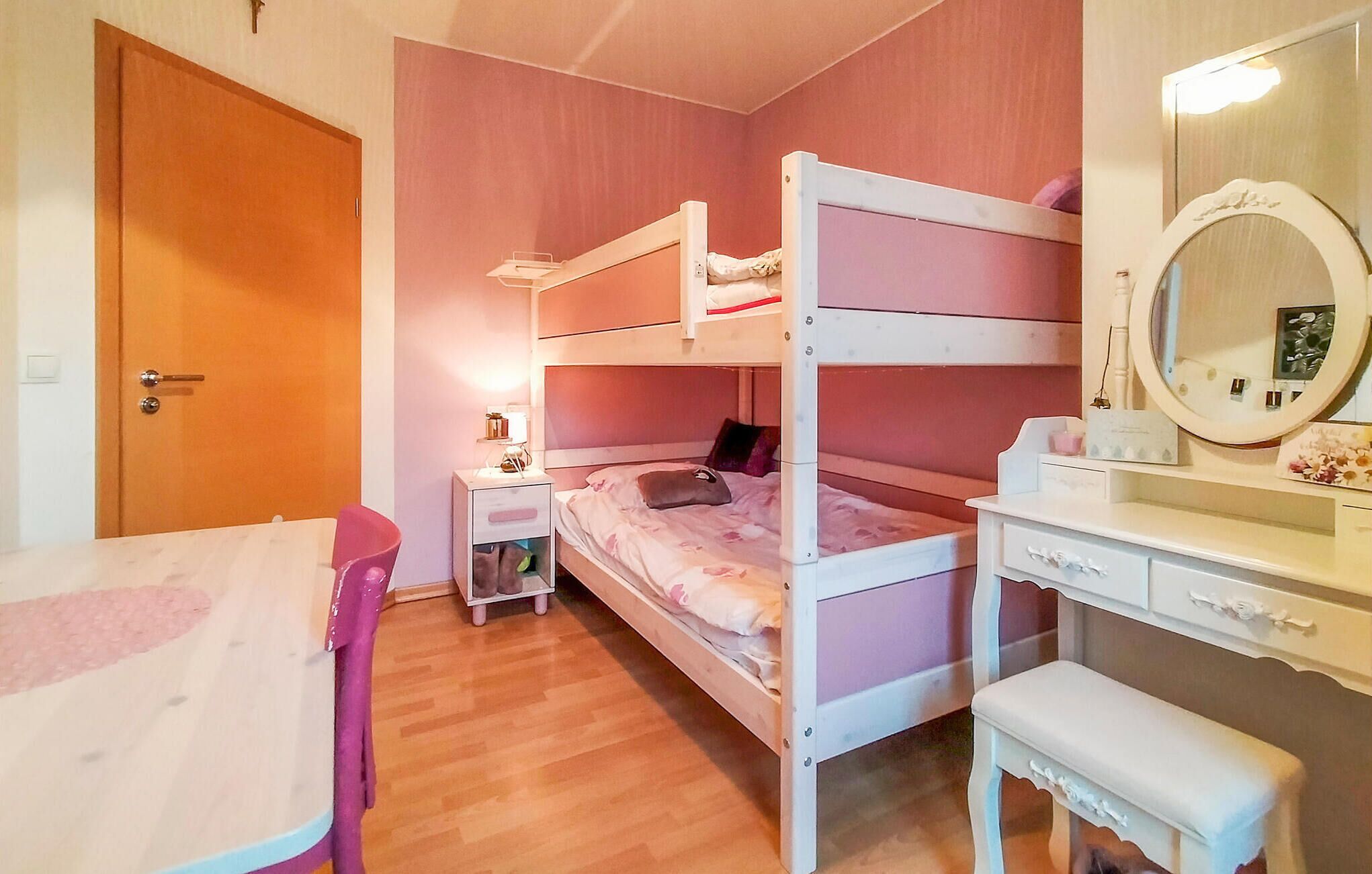 A bunk bed room with a wooden door, a bedside table with a lamp, a small chair, a vanity with a mirror, and a bench.
