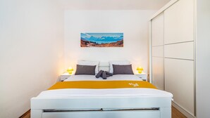 Ein Bett mit weißem Gestell, einer gelben Decke und zwei Kissen. Darüber an der Wand hängt ein Gemälde von Bergen.